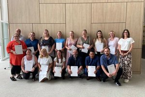 Gruppenfoto von 16 Absolventinnen und Absolventen nach dem erfolgreichen Abschluss einer Weiterbildung. Die Gruppe steht gemeinsam in den Räumlichkeiten der Zentralschule der Alexianer, dem Bildungszentrum für Gesundheit und Soziales. Die Teilnehmenden lächeln in die Kamera und halten gemeinsam den Abschluss ihrer Weiterbildung fest.