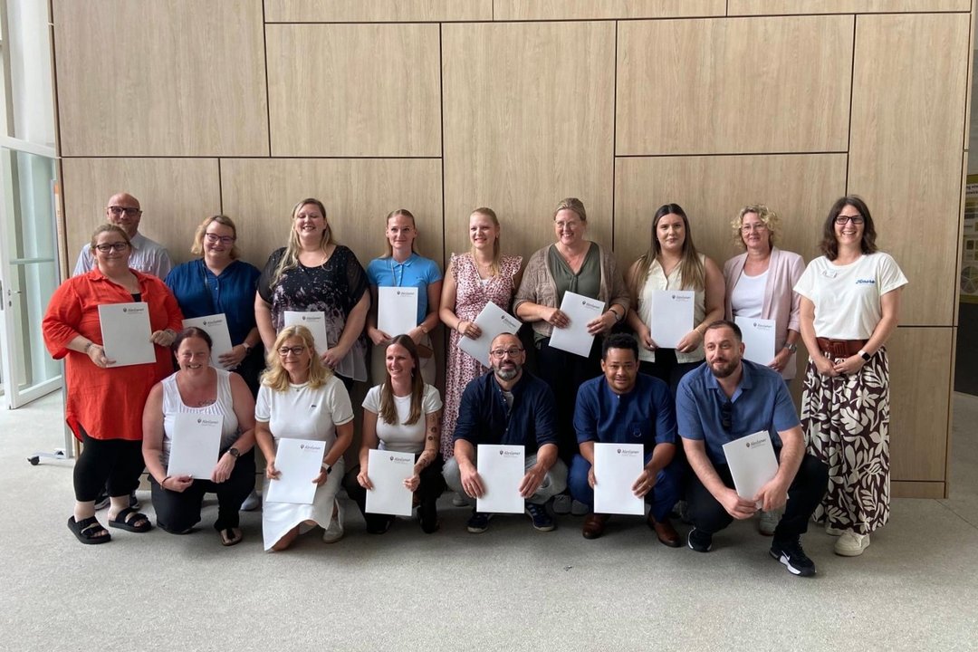 Gruppenfoto von 16 Absolventinnen und Absolventen nach dem erfolgreichen Abschluss einer Weiterbildung. Die Gruppe steht gemeinsam in den Räumlichkeiten der Zentralschule der Alexianer, dem Bildungszentrum für Gesundheit und Soziales. Die Teilnehmenden lächeln in die Kamera und halten gemeinsam den Abschluss ihrer Weiterbildung fest.